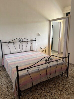 Cama com estrutura de metal e colcha listrada colorida em quarto com piso de azulejos e porta que dá para outro cômodo.