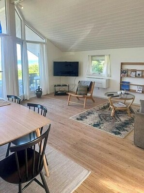 Ein geräumiges Wohnzimmer mit Holzboden, einem Esstisch mit Stühlen, einem Fernseher und einem großen Fenster.