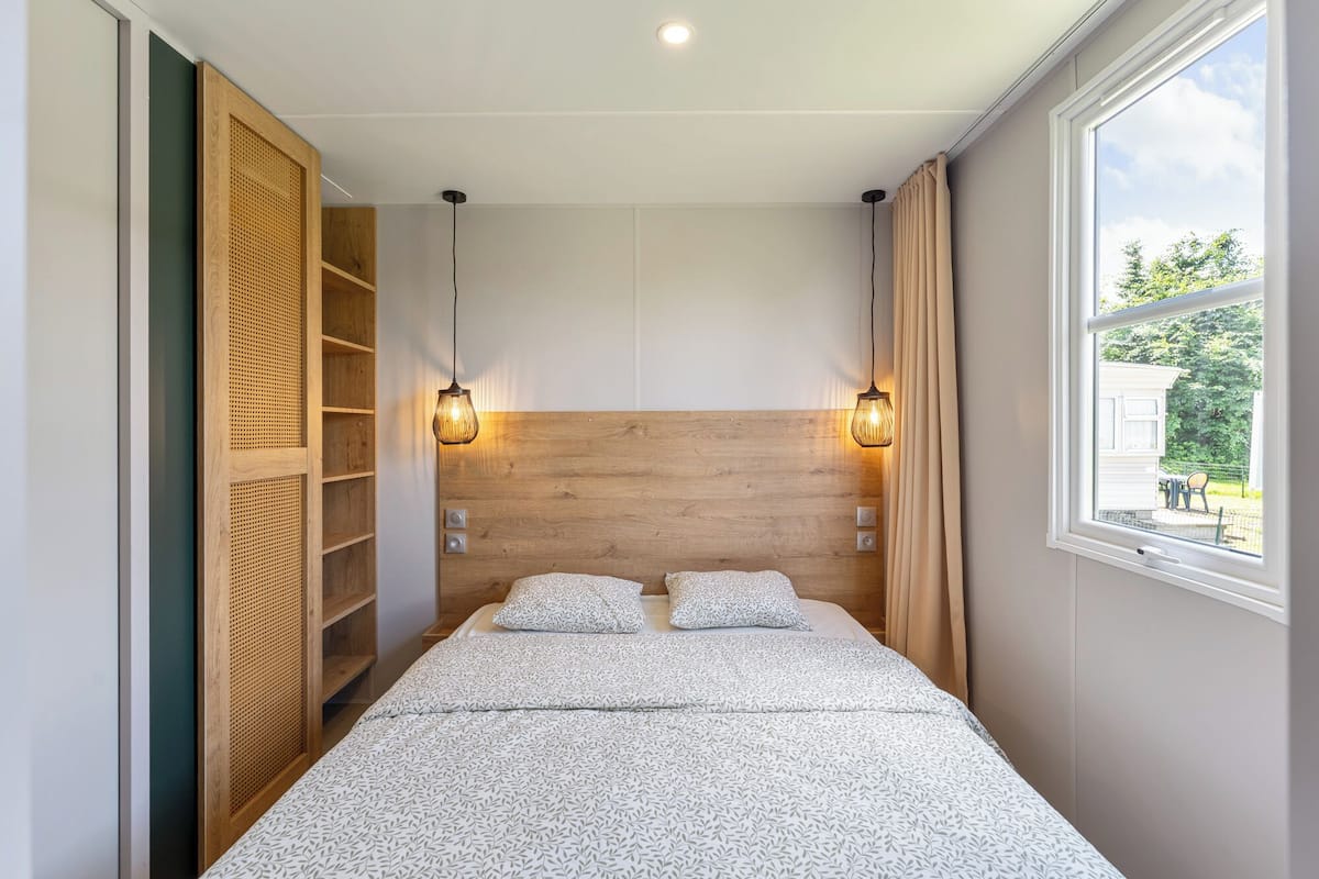 A bedroom with a wooden headboard, two bedside lamps, a window with curtains, and a bed with patterned bedding.