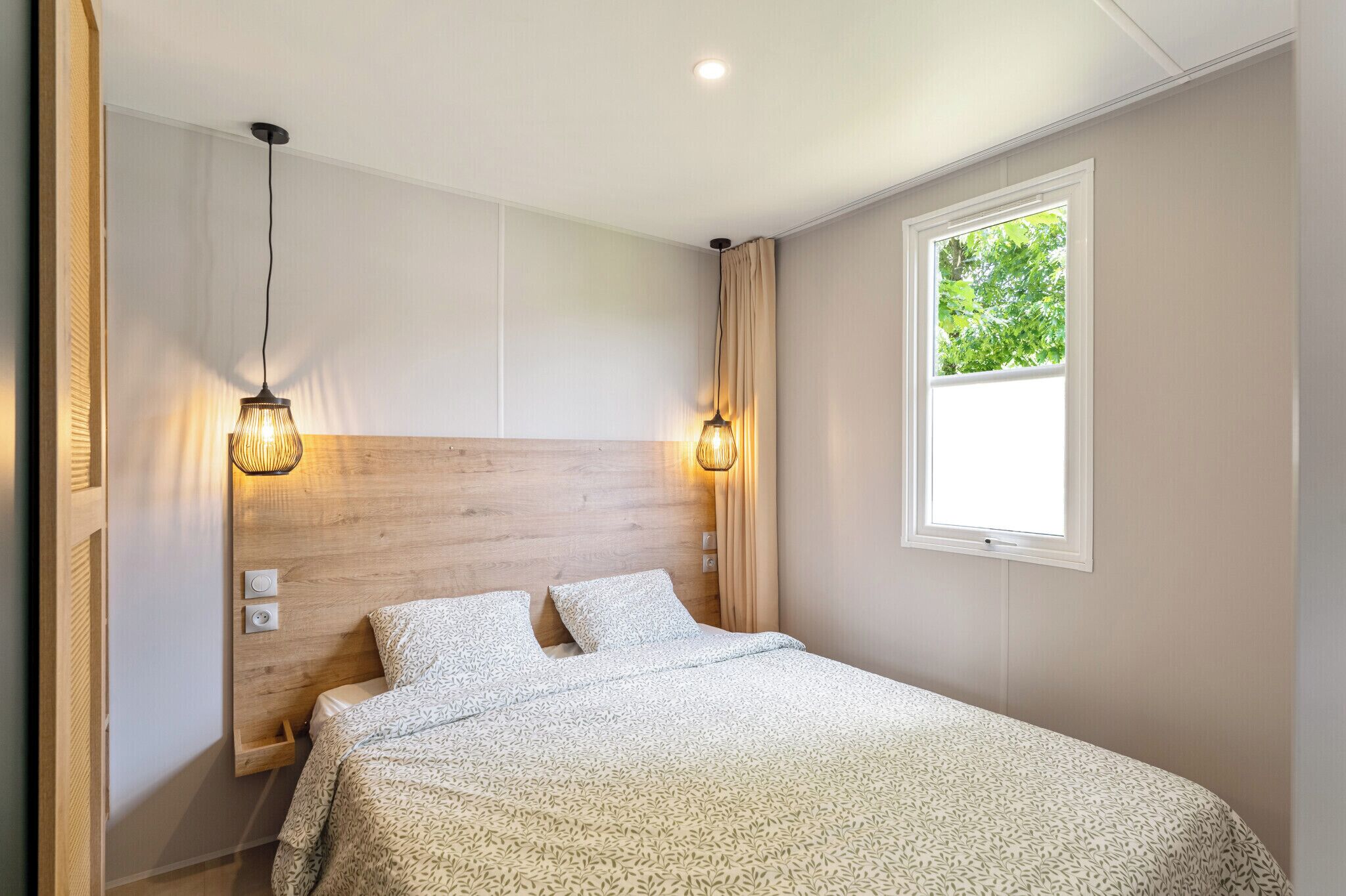 A bedroom with a wooden headboard, two bedside lamps, a window with a view of trees, and a bed with patterned bedding.