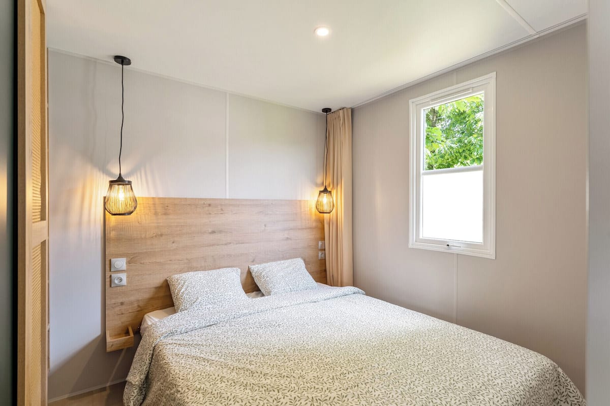 A bedroom with a wooden headboard, two bedside lamps, a window with a view of trees, and a bed with patterned bedding.