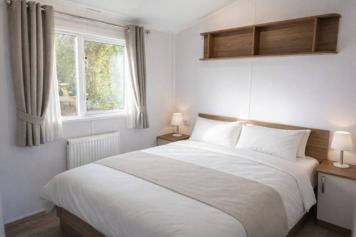 A bedroom with a bed, two bedside tables, a window with curtains, a radiator, and a wooden shelf above the bed.