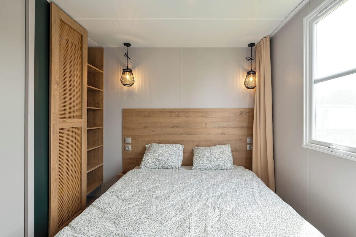 A bedroom with a wooden headboard, two bedside lamps, a bed with patterned bedding, and a window with curtains.