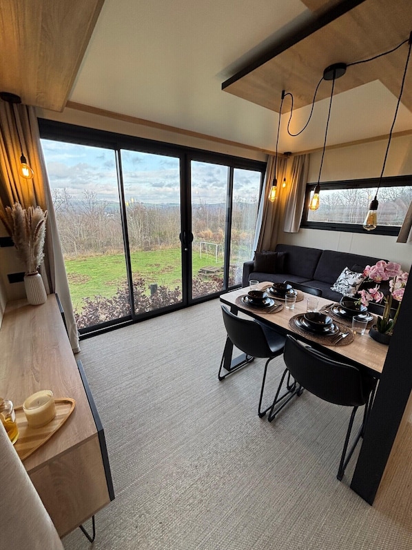 A modern dining area with a large window, a wooden table set for two, and a black sofa.