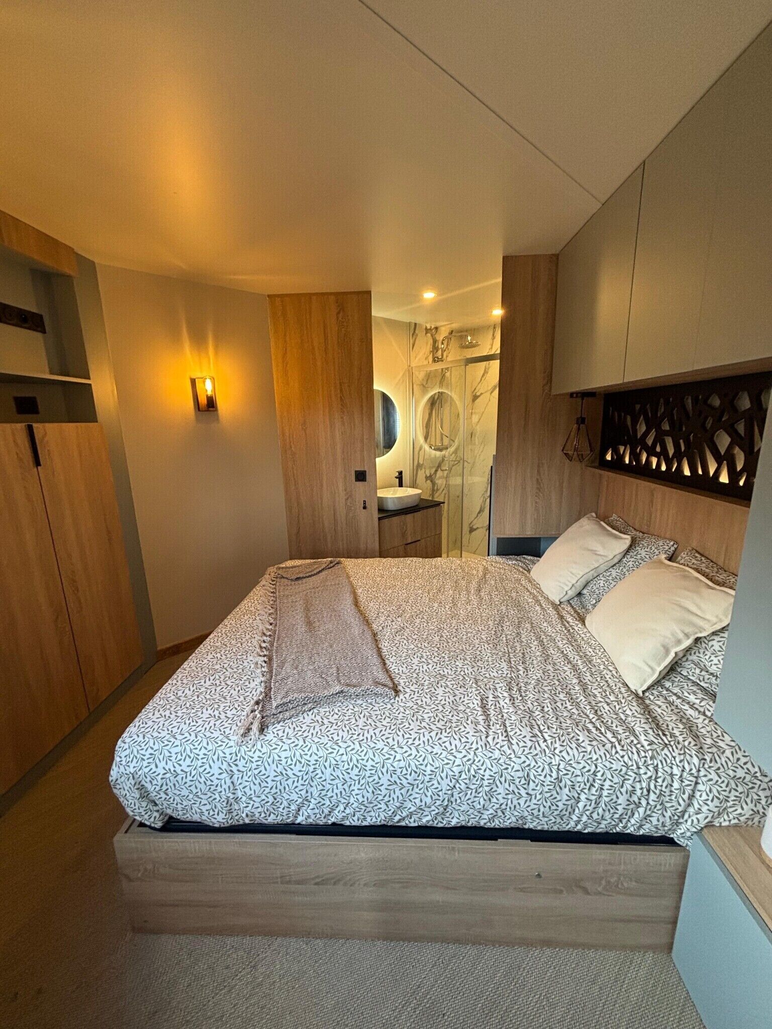 A modern bedroom with a bed, a wooden wardrobe, a bathroom with a marble shower, and a wall-mounted light fixture.