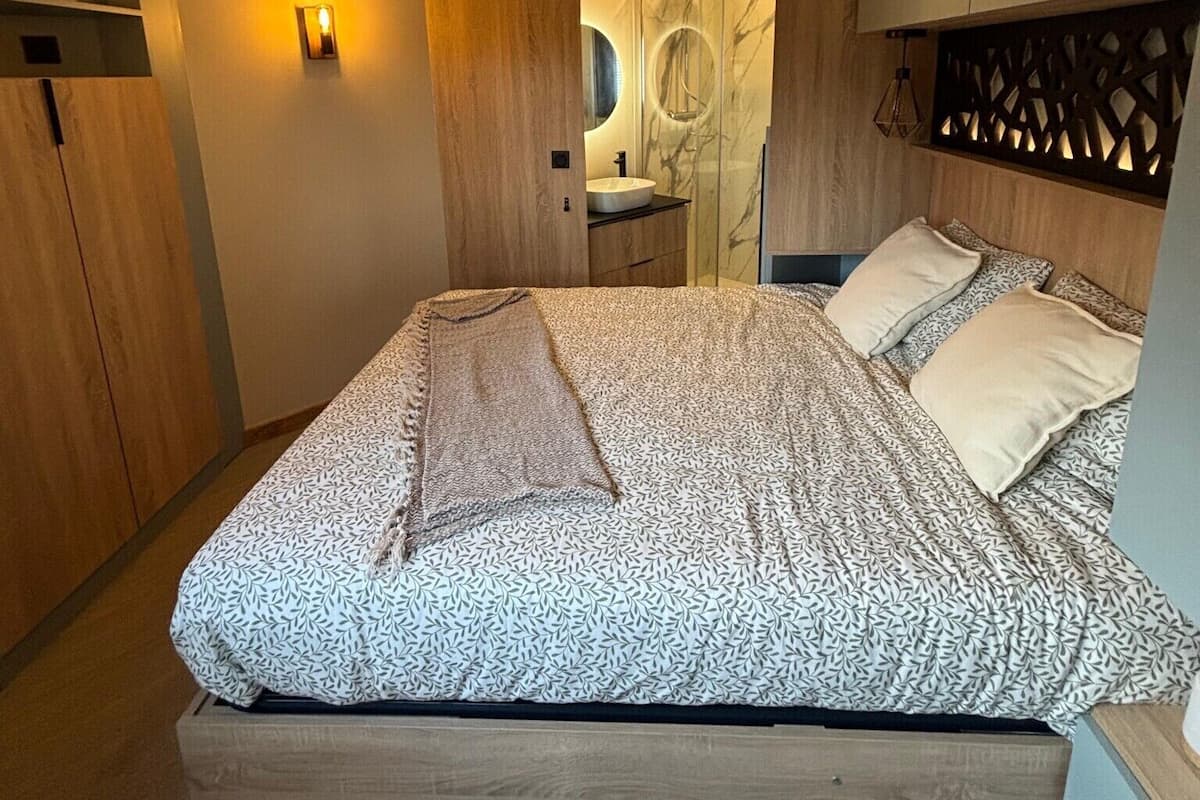 A modern bedroom with a bed, a wooden wardrobe, a bathroom with a marble shower, and a wall-mounted light fixture.