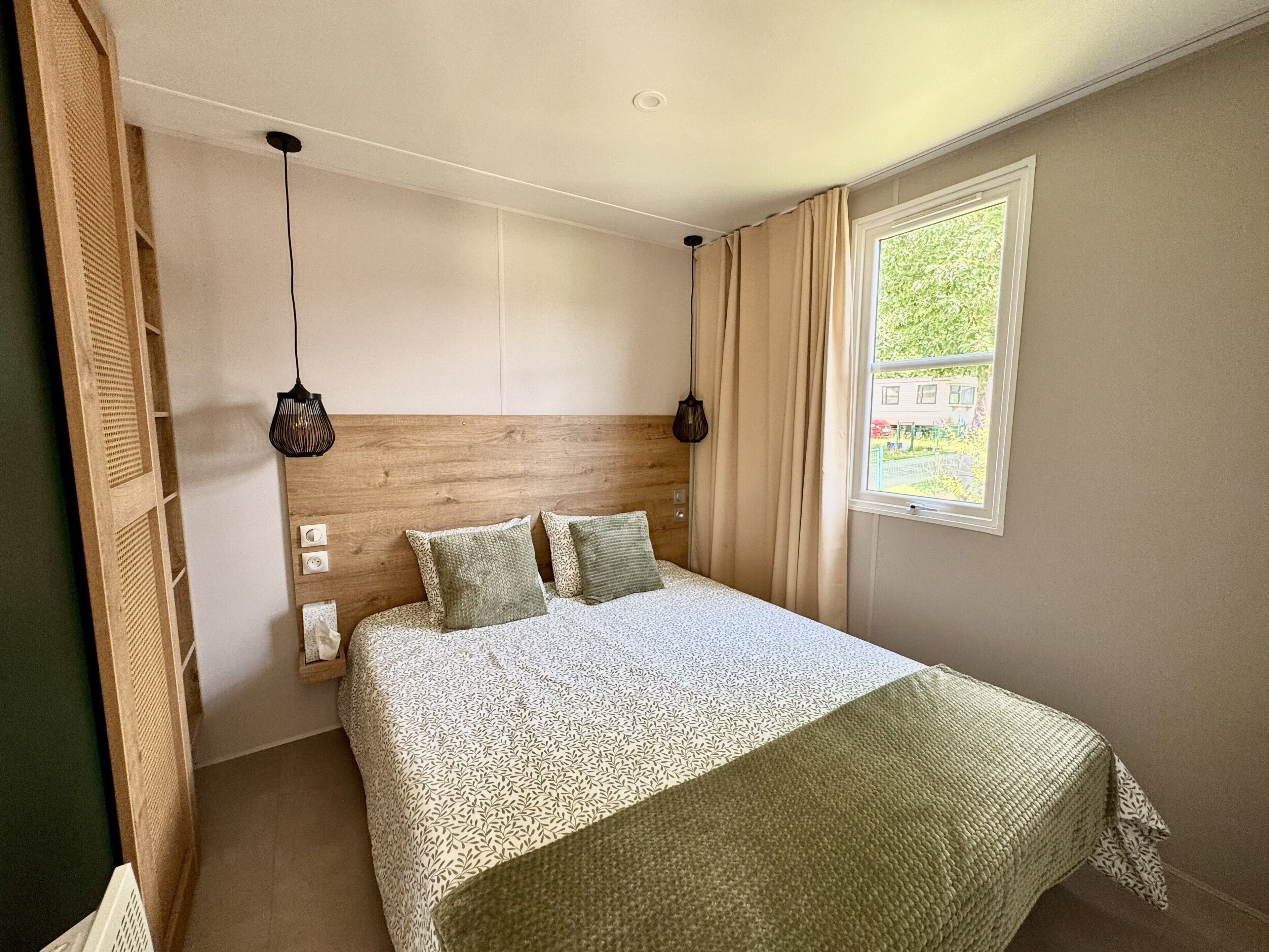 A bedroom with a wooden headboard, a patterned bedspread, two hanging lamps, and a window with curtains.