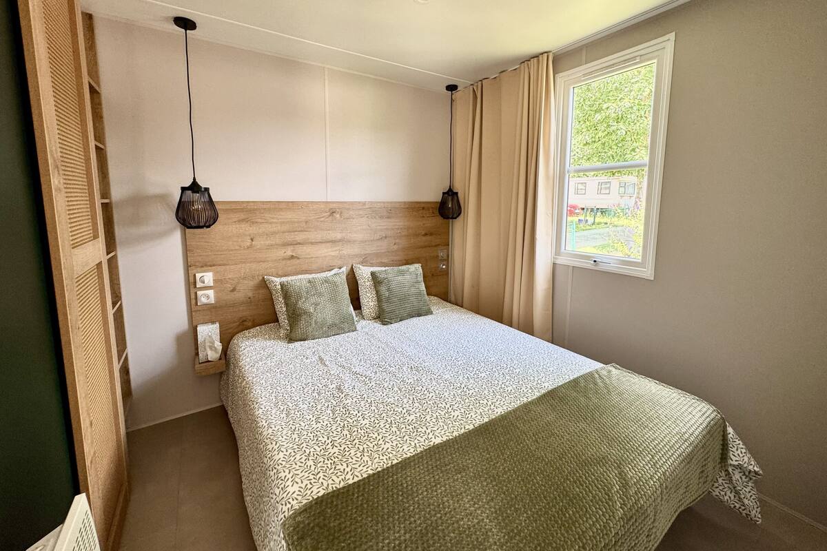 A bedroom with a wooden headboard, a patterned bedspread, two hanging lamps, and a window with curtains.