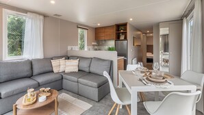 Una sala de estar moderna con un sofá seccional gris, una mesa de comedor con sillas blancas y una cocina con armarios de madera y un refrigerador.