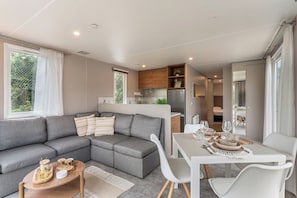 A modern living room with a grey sectional sofa, a dining table with white chairs, and a kitchen area with wooden cabinets and a refrigerator.