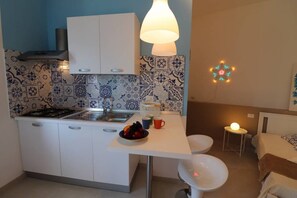 Une cuisine compacte équipée d’une cuisinière à gaz, d’un évier et de meubles blancs. On y trouve une petite table à manger avec un saladier de fruits et des tasses. Un luminaire décoratif est suspendu au-dessus de la table.