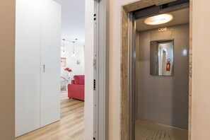 Een lift met een brandblusser, een spiegel en een deur die leidt naar een kamer met een rode stoel en kunstwerken.
