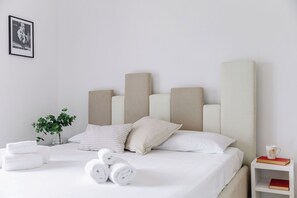 Camera d'albergo con un letto, cuscini, asciugamani arrotolati, una pianta sul comodino e un piccolo tavolo da notte con un libro e una tazza.