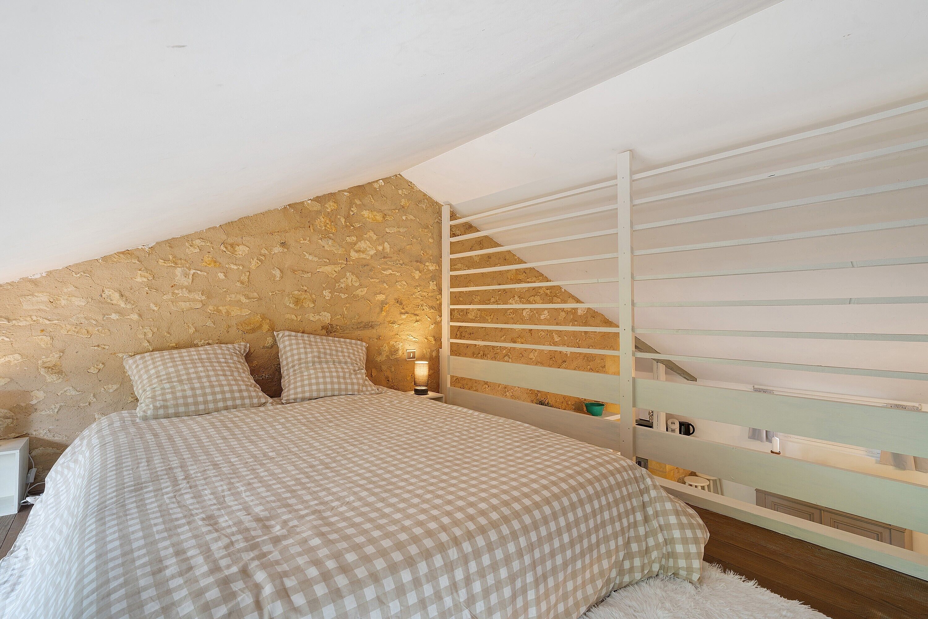 Une chambre à coucher avec une couvre-lit à carreaux, un mur en pierre, une balustrade blanche et une table de chevet avec une lampe.