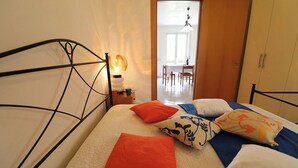 A bed with a quilted mattress, a bedside table with a lamp, and a view of a dining area through an open door. - Wonderful  villa for 4 guests with A/C, WIFI, TV and balcony (Otranto)