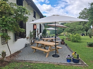 Une terrasse avec des tables et des bancs en bois, entourée de gravier, de plantes en pot et d’un bâtiment blanc aux volets noirs.