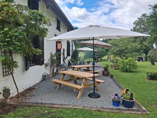Un patio con mesas y bancos de madera, rodeado de grava, plantas en macetas y un edificio blanco con persianas negras.
