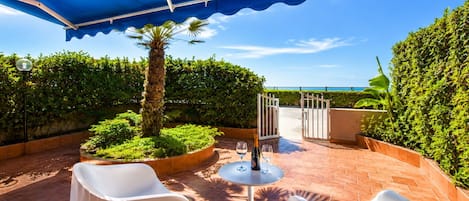 Eine Terrassengaststätte mit blauem Baldachin, weißen Stühlen, einem Tisch mit Weingläsern und Blick aufs Meer.