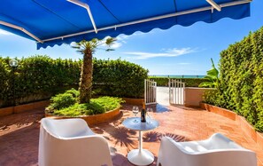Eine Terrassengaststätte mit blauem Baldachin, weißen Stühlen, einem Tisch mit Weingläsern und Blick aufs Meer.