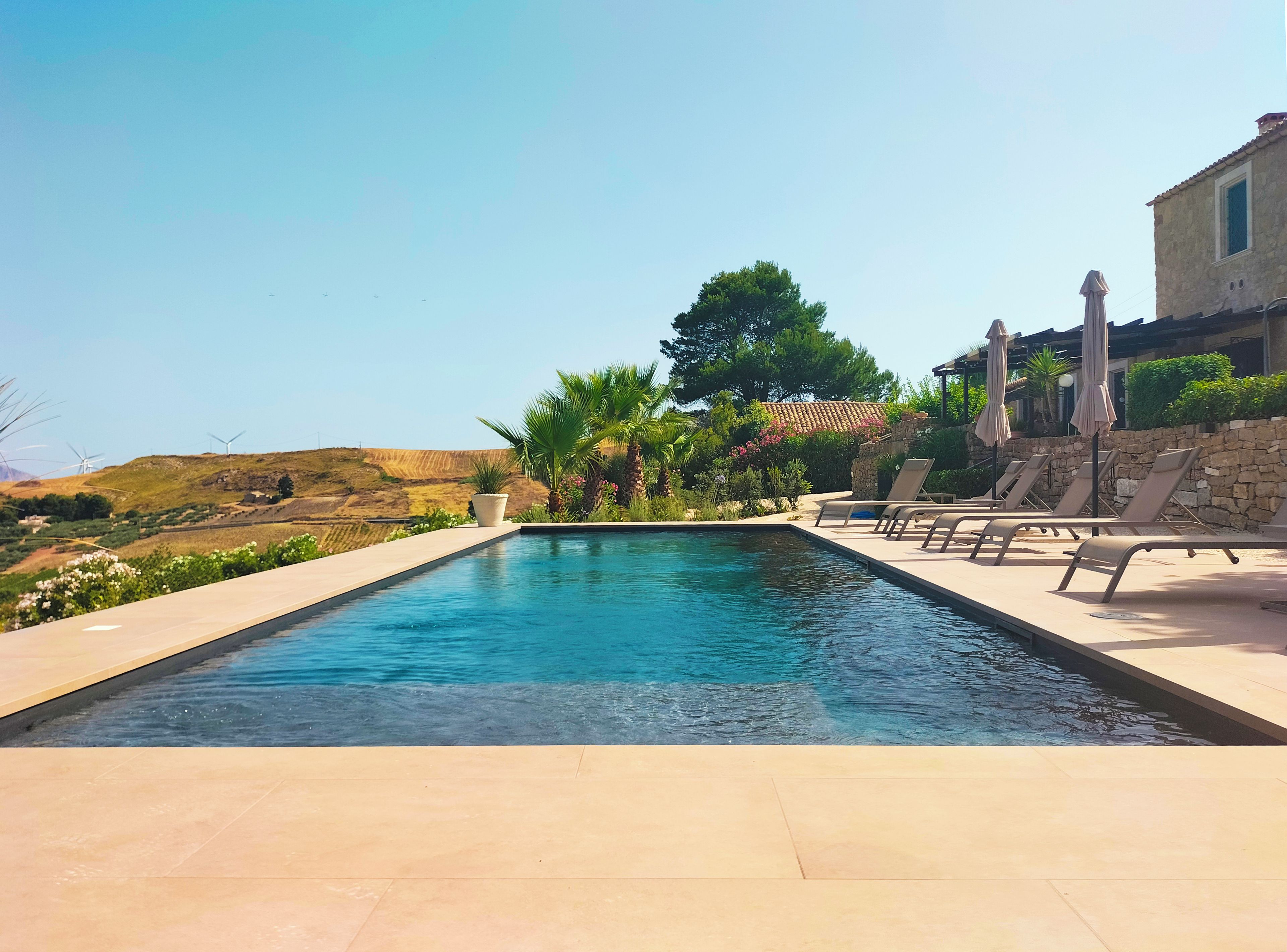 A swimming pool with lounge chairs and umbrellas, surrounded by greenery and a stone building.