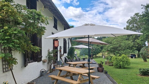 Une terrasse avec des tables et des bancs en bois, entourée de gravier, de plantes en pot et d’un bâtiment blanc aux volets noirs.