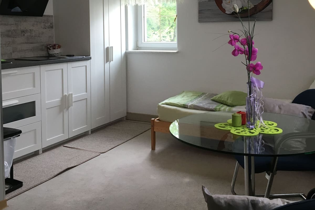 A compact kitchen with white cabinets, a window with sheer curtains, a dining area with a glass table, and a painting on the wall.