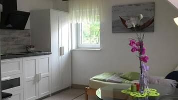 Cocina compacta con armarios blancos, una ventana con cortinas traslúcidas, un comedor con mesa de vidrio y un cuadro en la pared.