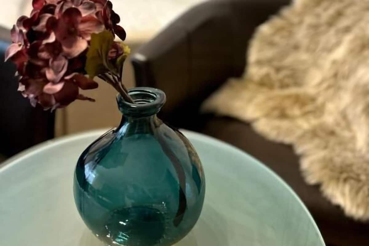 A teal vase with flowers on a table, a bed with white bedding, and a fur throw on a couch.