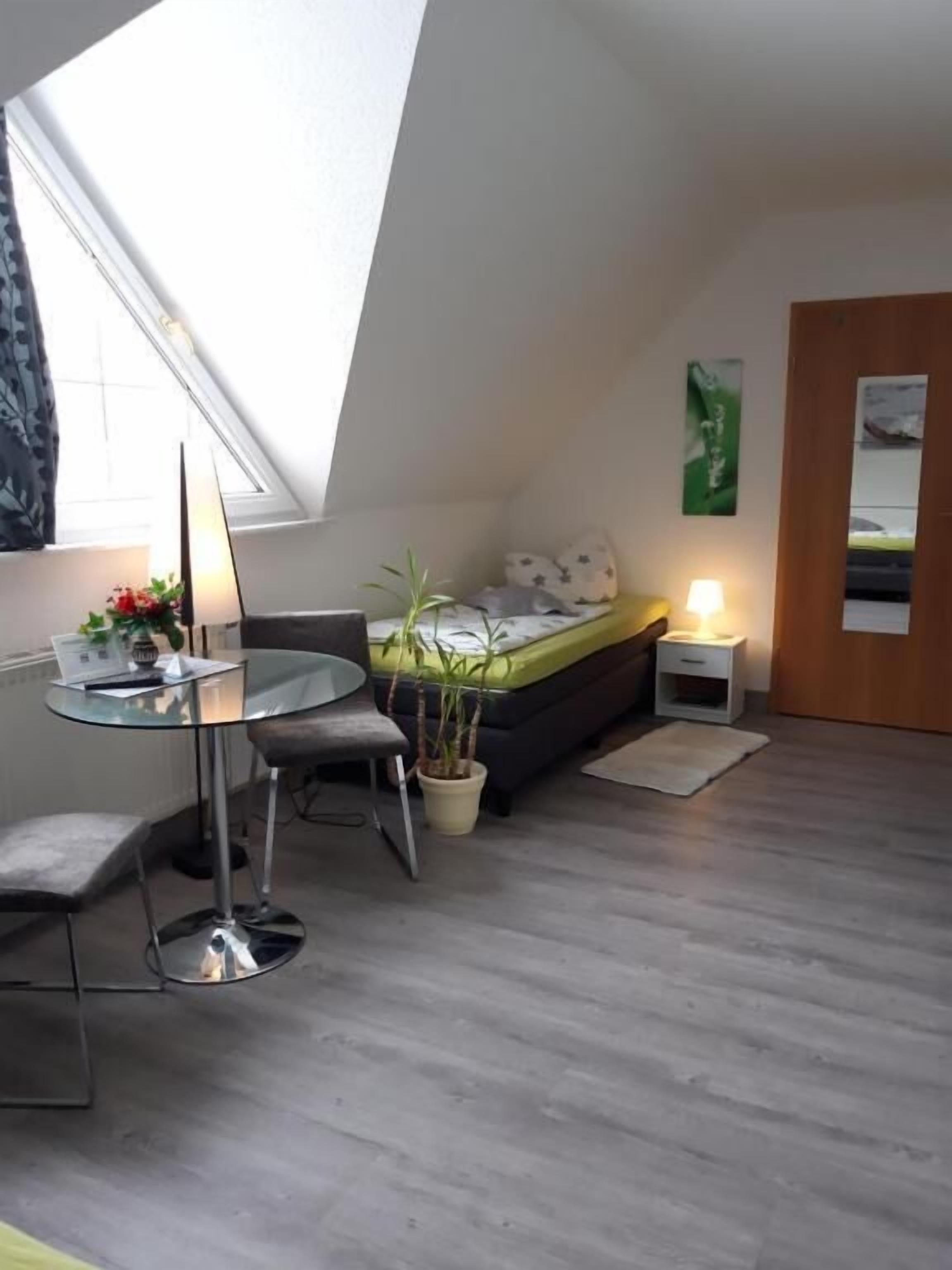 Ein kleiner, moderner Raum mit Dachfenster, einem runden Tisch, einem Bett, einem Nachttisch und einer Topfpflanze.