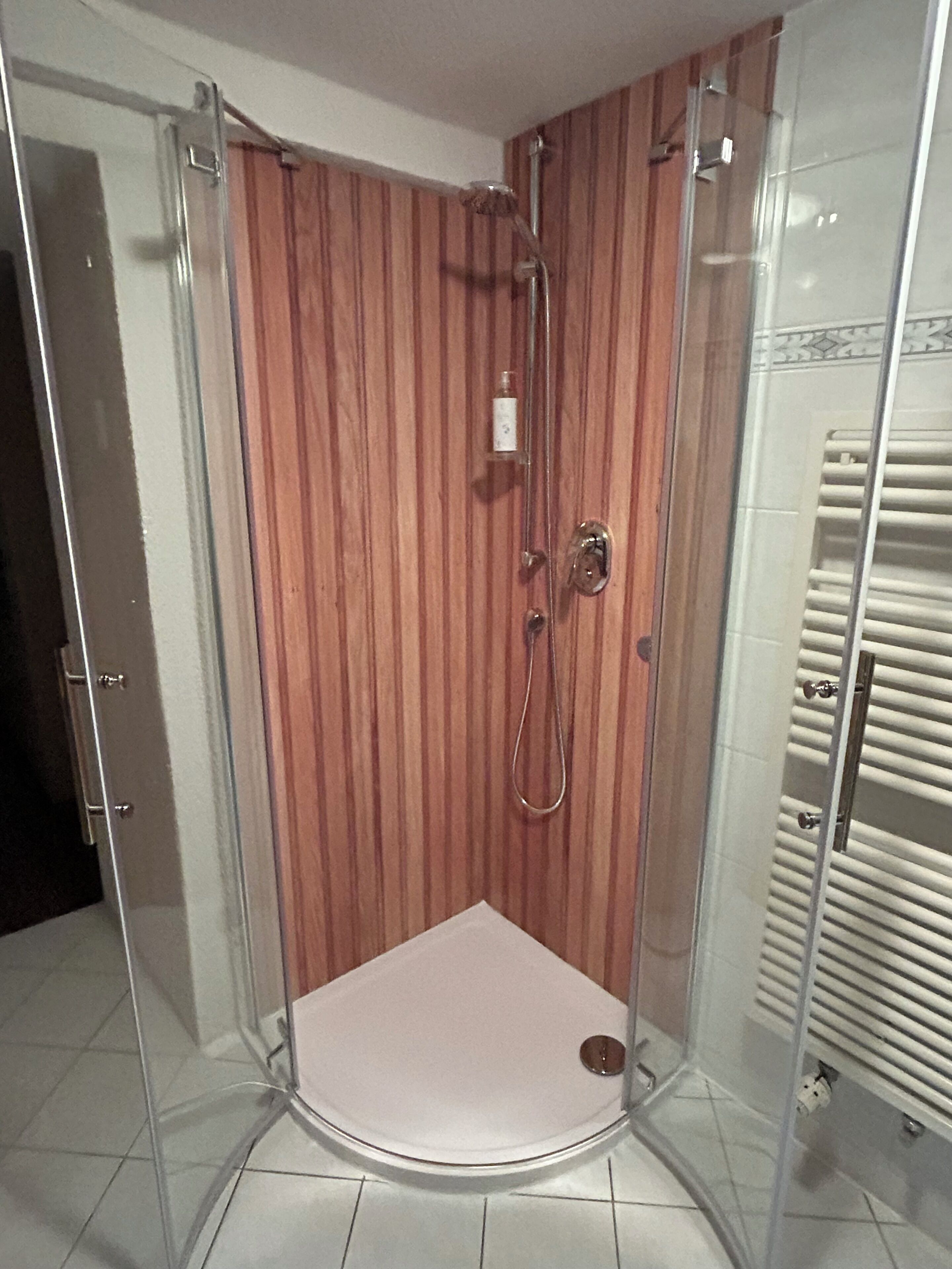 A glass-enclosed shower stall with a wooden panel wall, a white seat, and a visible showerhead.