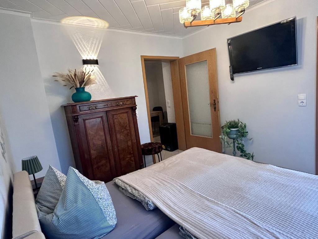A bedroom with a bed, a wooden dresser, a TV, and a door leading to another room.