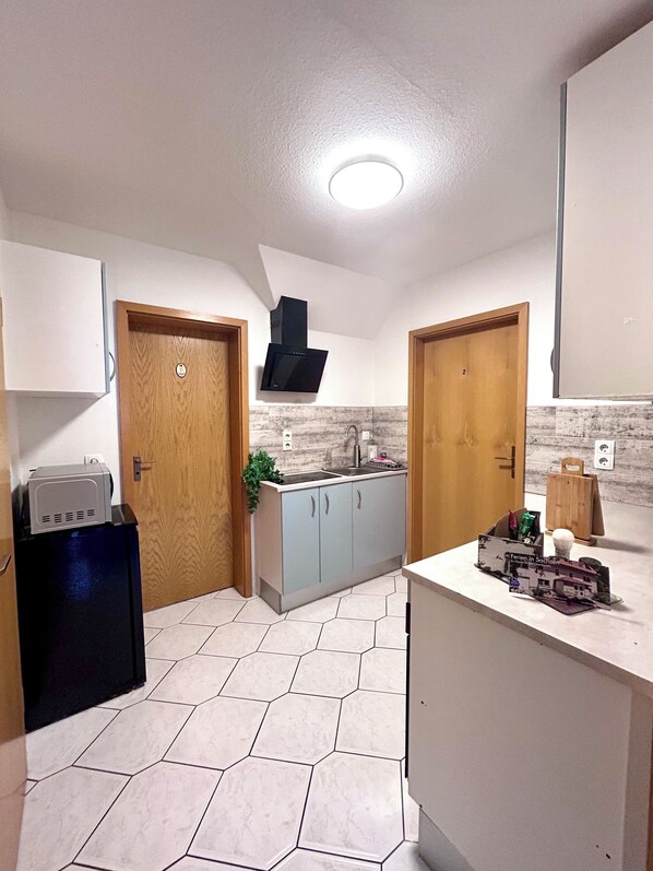 Cocina compacta con estufa integrada, pileta y campana extractora negra. Cuenta con dos puertas de madera, un refrigerador pequeño y una mesada con un objeto decorativo.