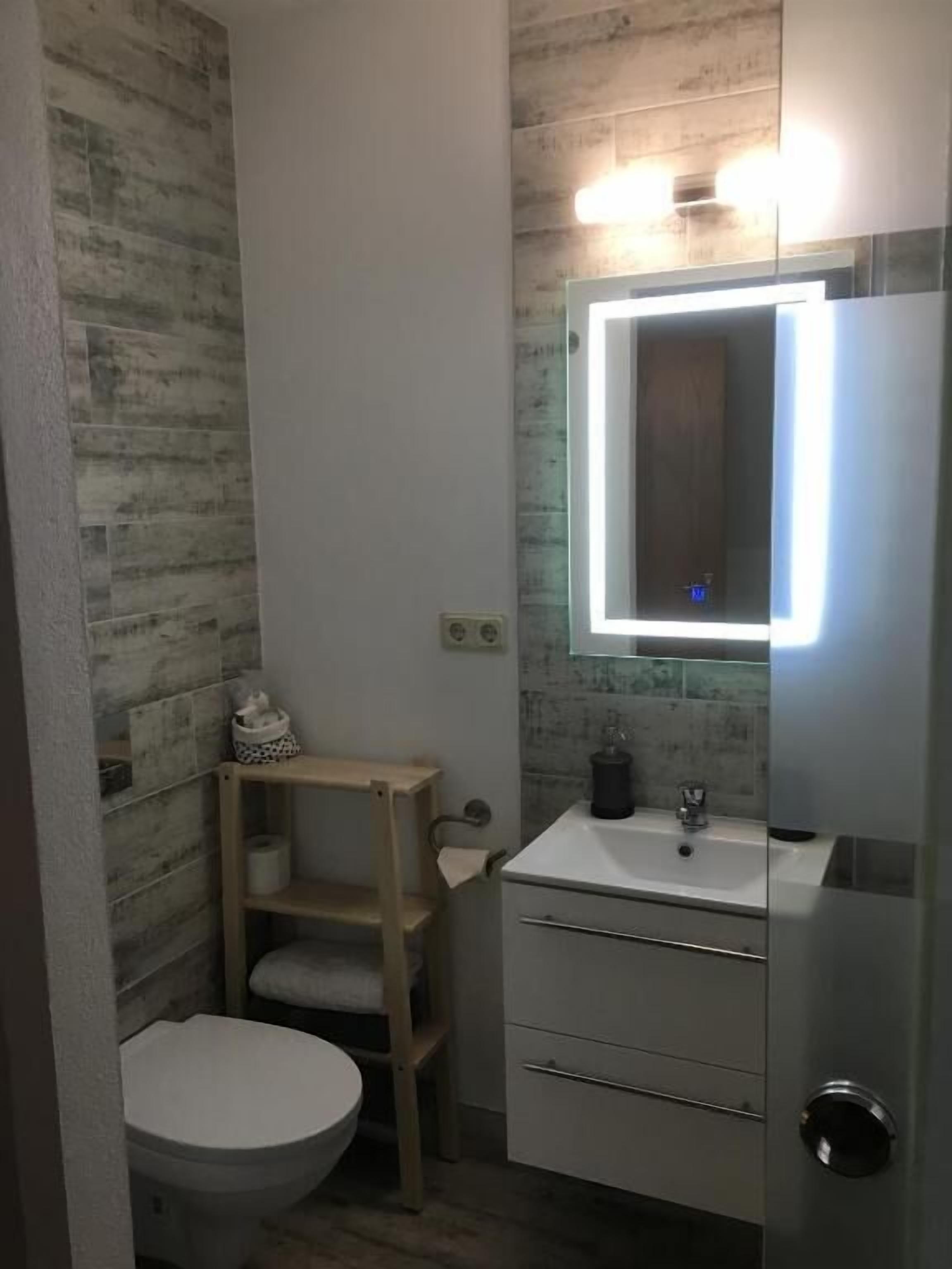Une salle de bain comprenant des toilettes, une étagère en bois, un meuble-lavabo avec un miroir et un évier.