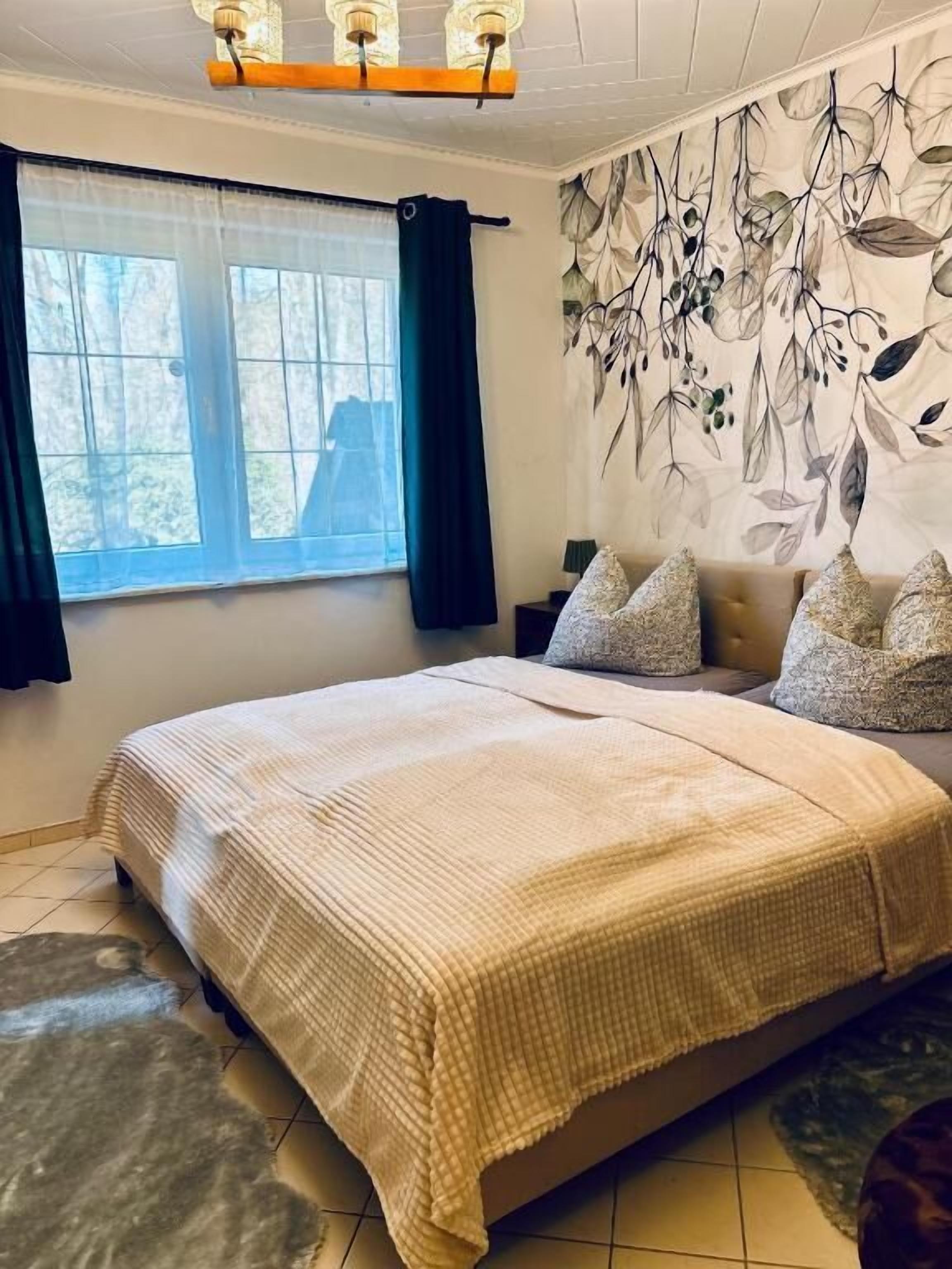 A bedroom with a bed, a window with curtains, a wall with a leaf pattern, and a ceiling light fixture.
