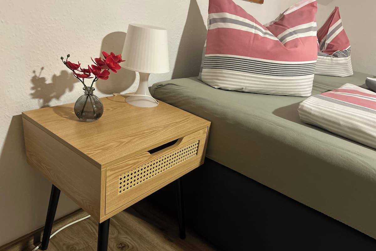 A neatly arranged bedroom with a wooden nightstand, a lamp, a vase with flowers, and a bed with striped pillows.