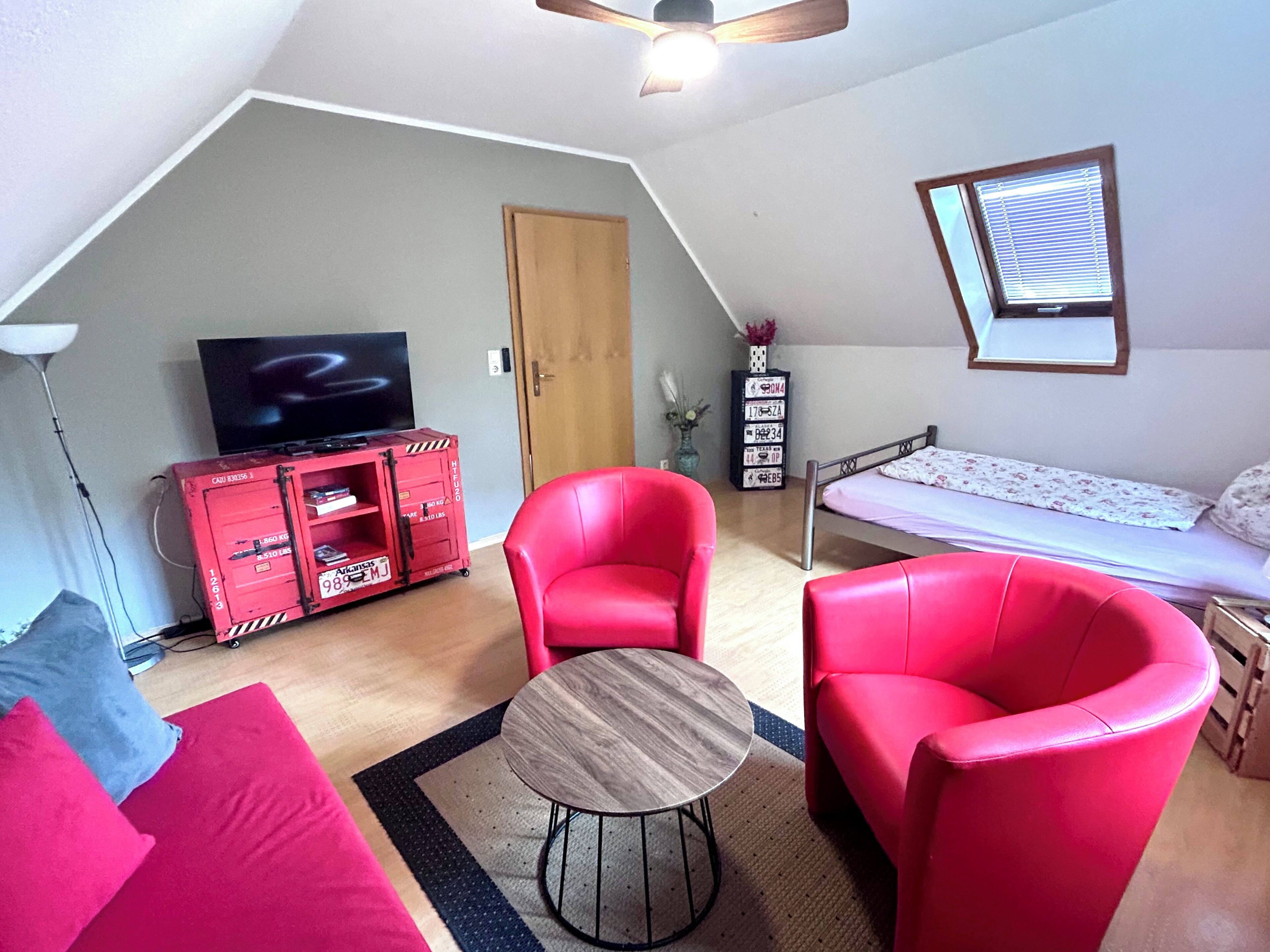 A room with a loft bed, a red sofa, a red armchair, a small round table, a television on a red cabinet, and a window with blinds.