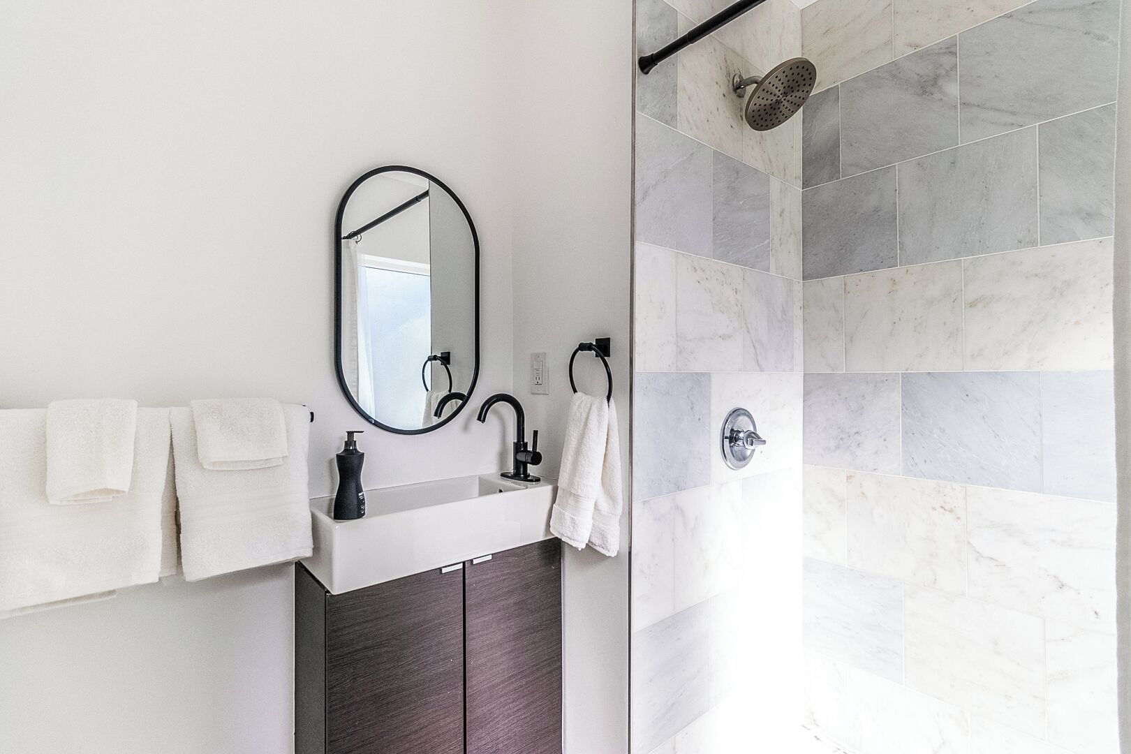 Un baño moderno con un lavamanos blanco, un mueble de madera oscura, un espejo redondo y una ducha con puerta de vidrio.