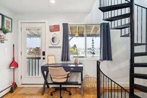 Une pièce comprenant un bureau, une chaise, une fenêtre avec des rideaux, une serpillière rouge, un panier en osier et un escalier en colimaçon noir.