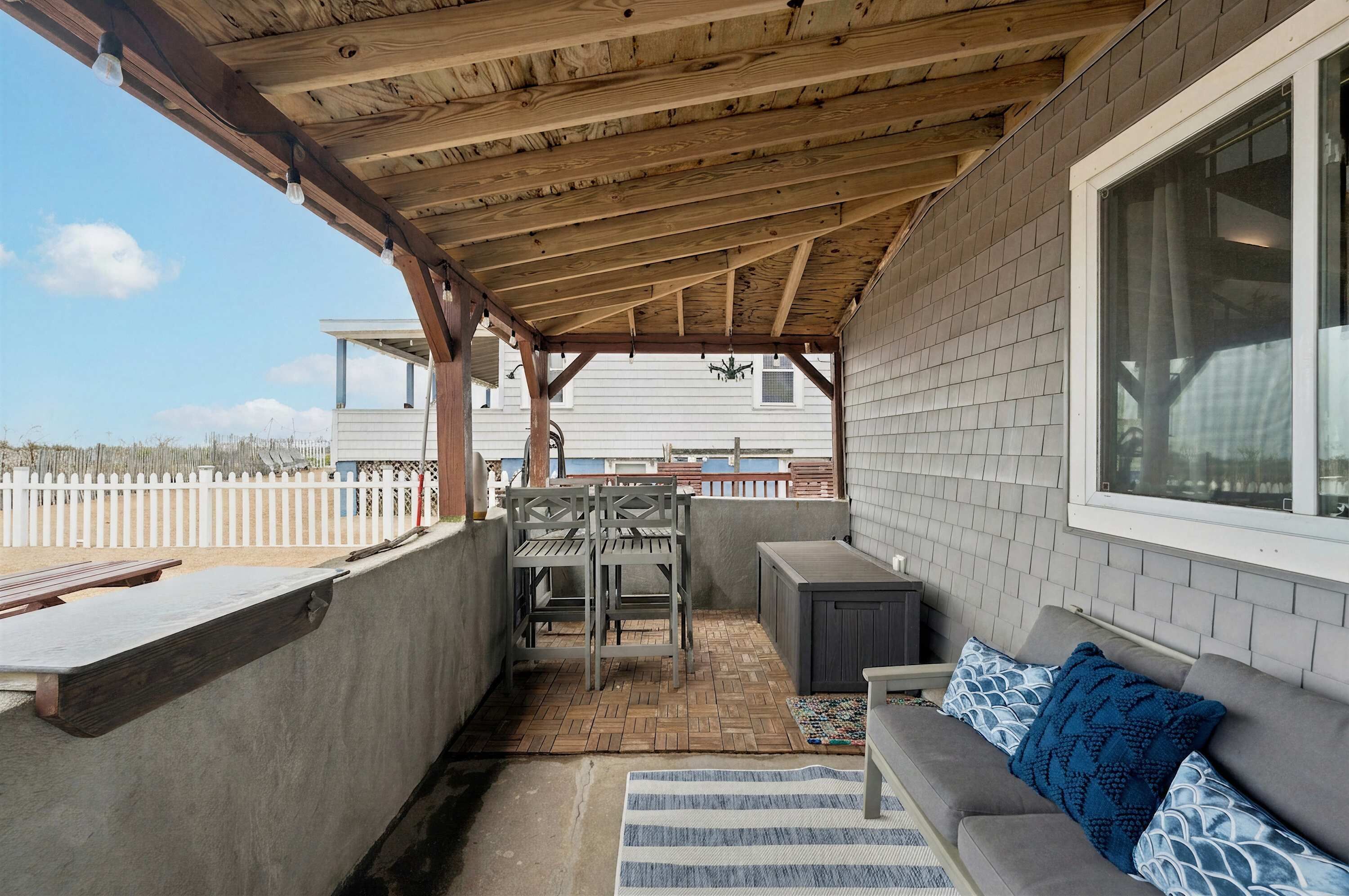Une terrasse couverte avec un plafond en bois, un canapé gris agrémenté de coussins à motifs, une petite table et un banc.