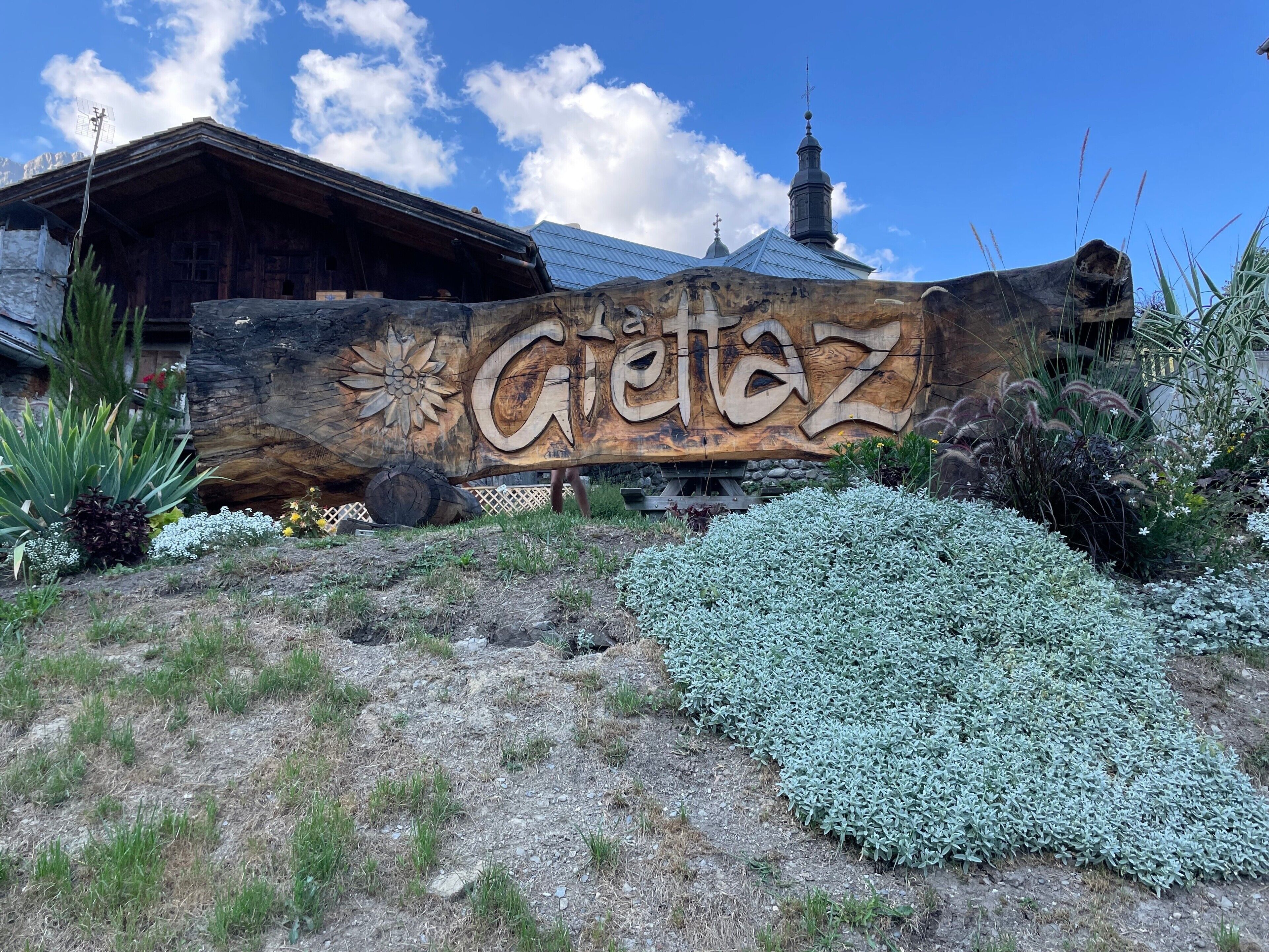 Ein hölzernes Schild mit der eingravierten Aufschrift 'Gretz', umgeben von einem angelegten Garten mit verschiedenen Pflanzen.