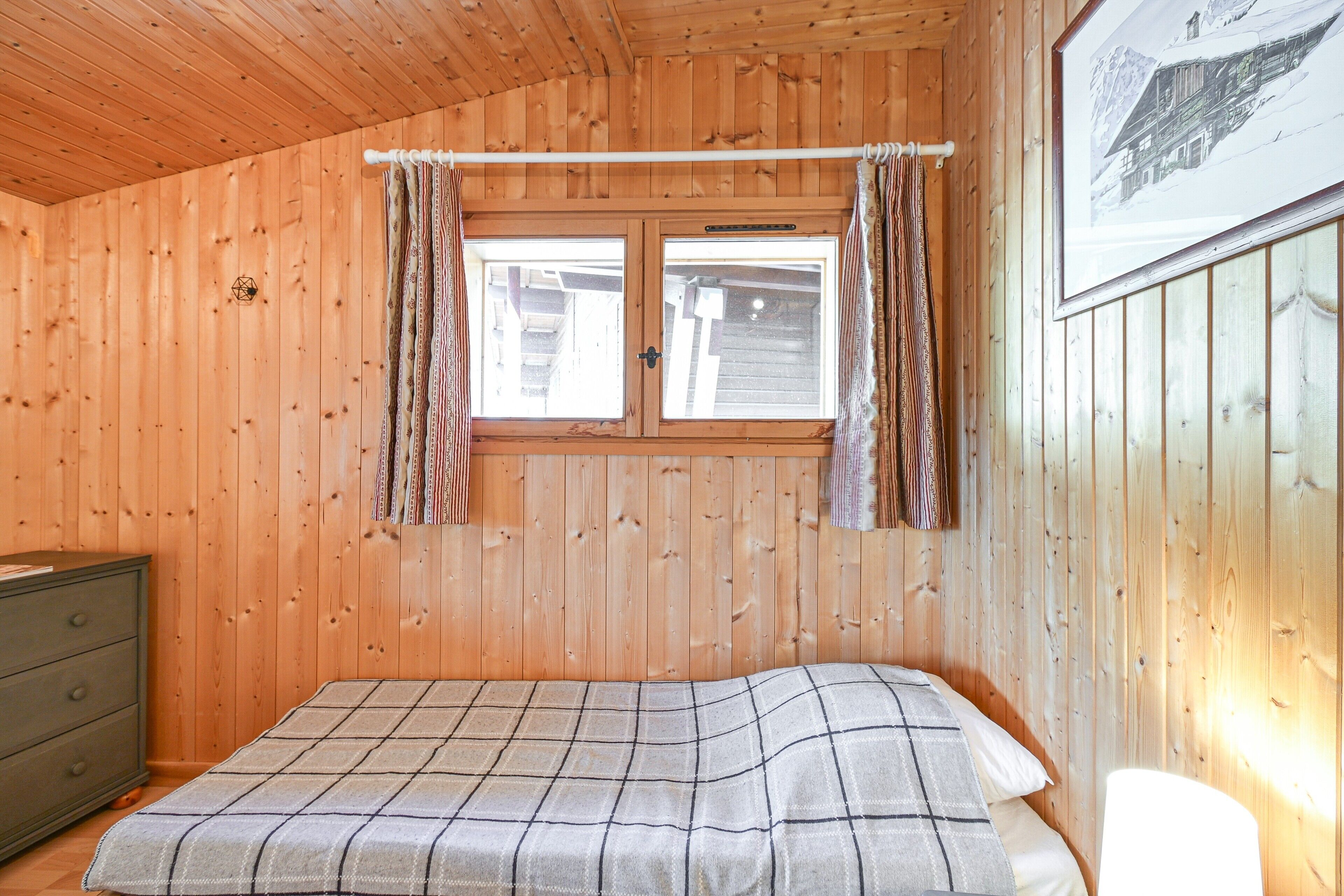 Une pièce de cabane en bois comprenant un lit, une commode et une fenêtre avec des rideaux.