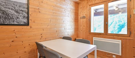 Interior de una cabaña de madera con una mesa blanca, dos sillas, un radiador y una ventana con vista a los árboles.