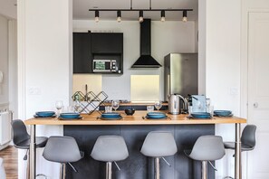 A modern kitchen with a central island, bar stools, a microwave, and a kettle. - Le Jeanne d’Arc – City center & sleeps 8 (Reims)