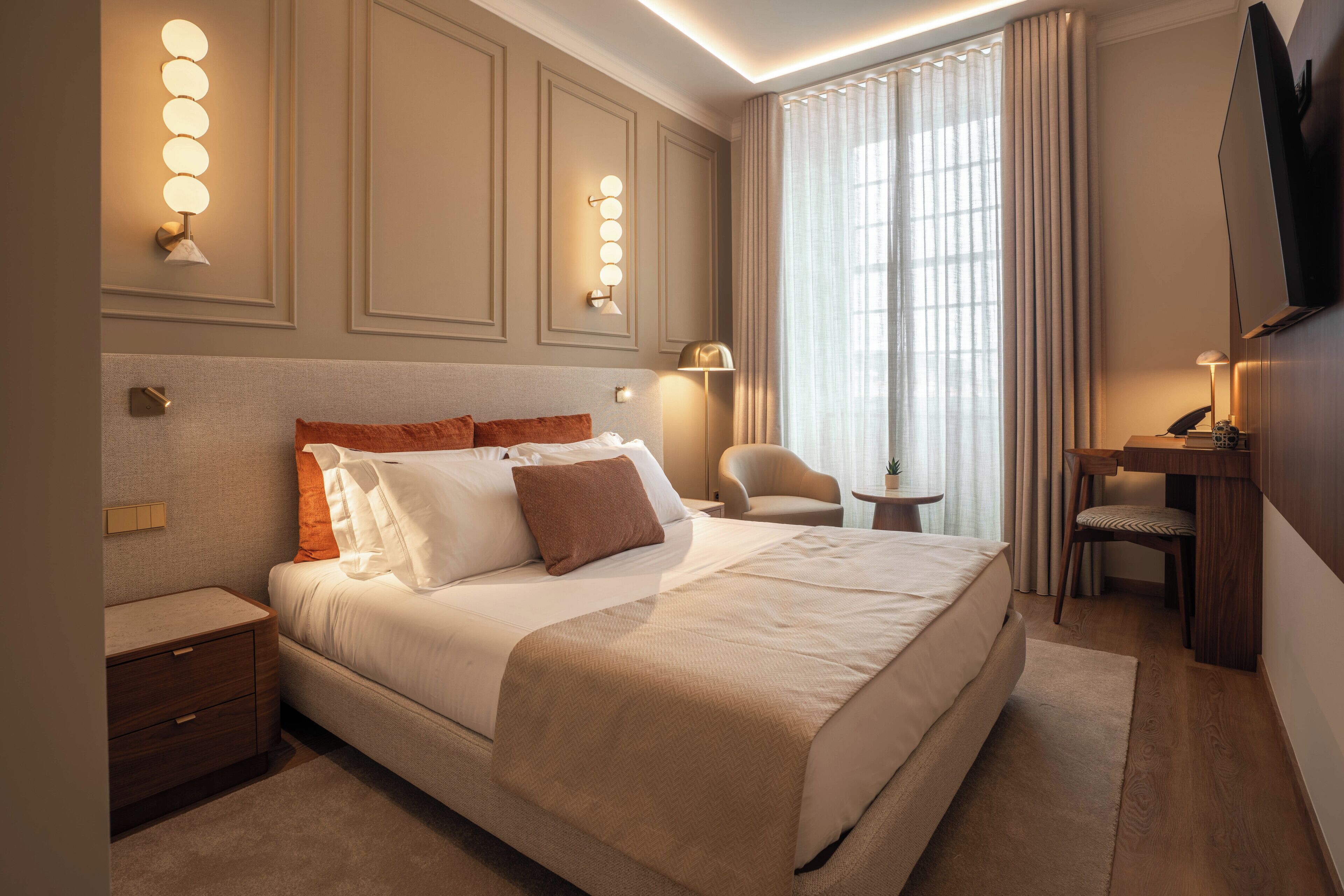 A hotel room with a large bed, bedside tables, a desk with a chair, a TV, and wall-mounted lighting.