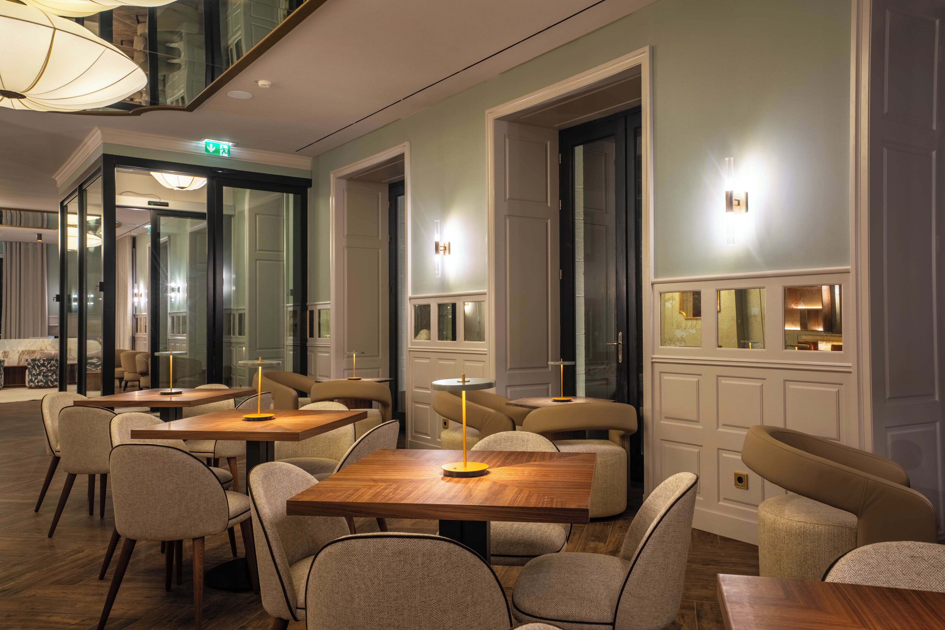 A modern hotel lobby with wooden tables and chairs, a glass partition, and a staircase.