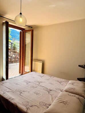 Ein Schlafzimmer mit Bett, einer Pendelleuchte, einer Heizung und Blick auf eine gebirgige Landschaft durch eine offene Tür.