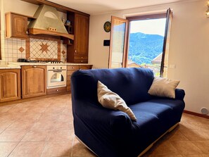 Ein Wohnzimmer mit blauem Sofa, Holzsärchen, einem Herd und einem Fenster mit Blick auf die Berge.
