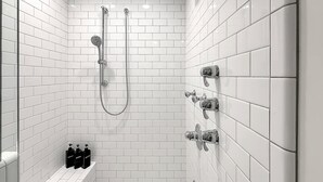 A modern bathroom with white tiled walls, a shower with a handheld showerhead, and a bench with bottles.