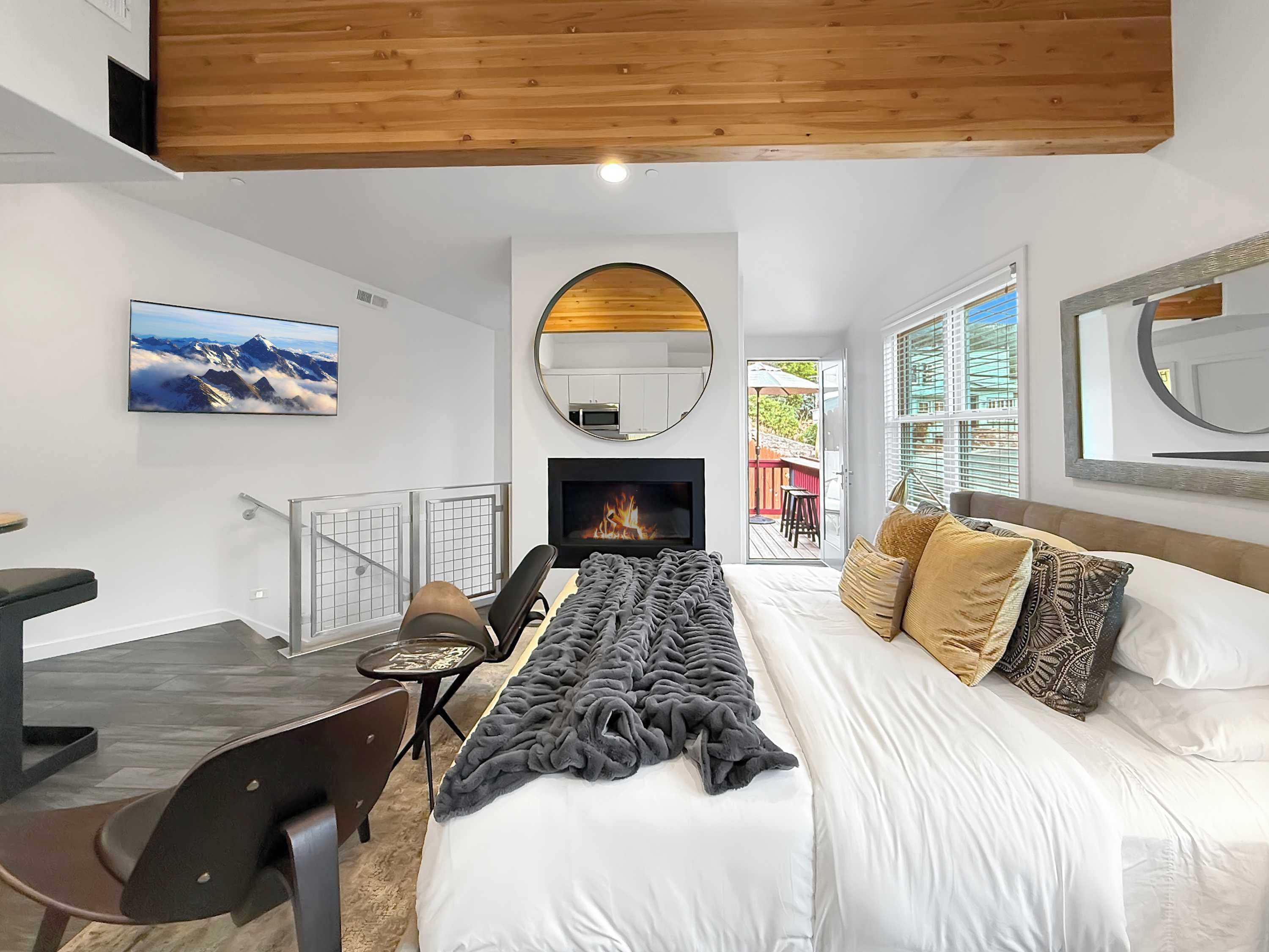A modern bedroom with a bed, a desk, a chair, a fireplace, and a large window.