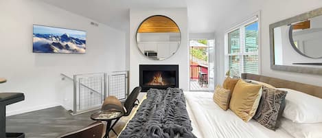 A modern bedroom with a bed, a desk, a chair, a fireplace, and a large window.